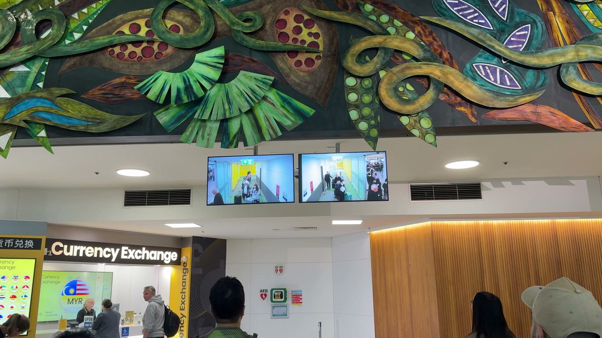 Ankunftsbereich am Flughafen Brisbane: zwei Monitore unter der Decke zeigen das Kamerabild des Gangs, durch den die Ankommenden laufen