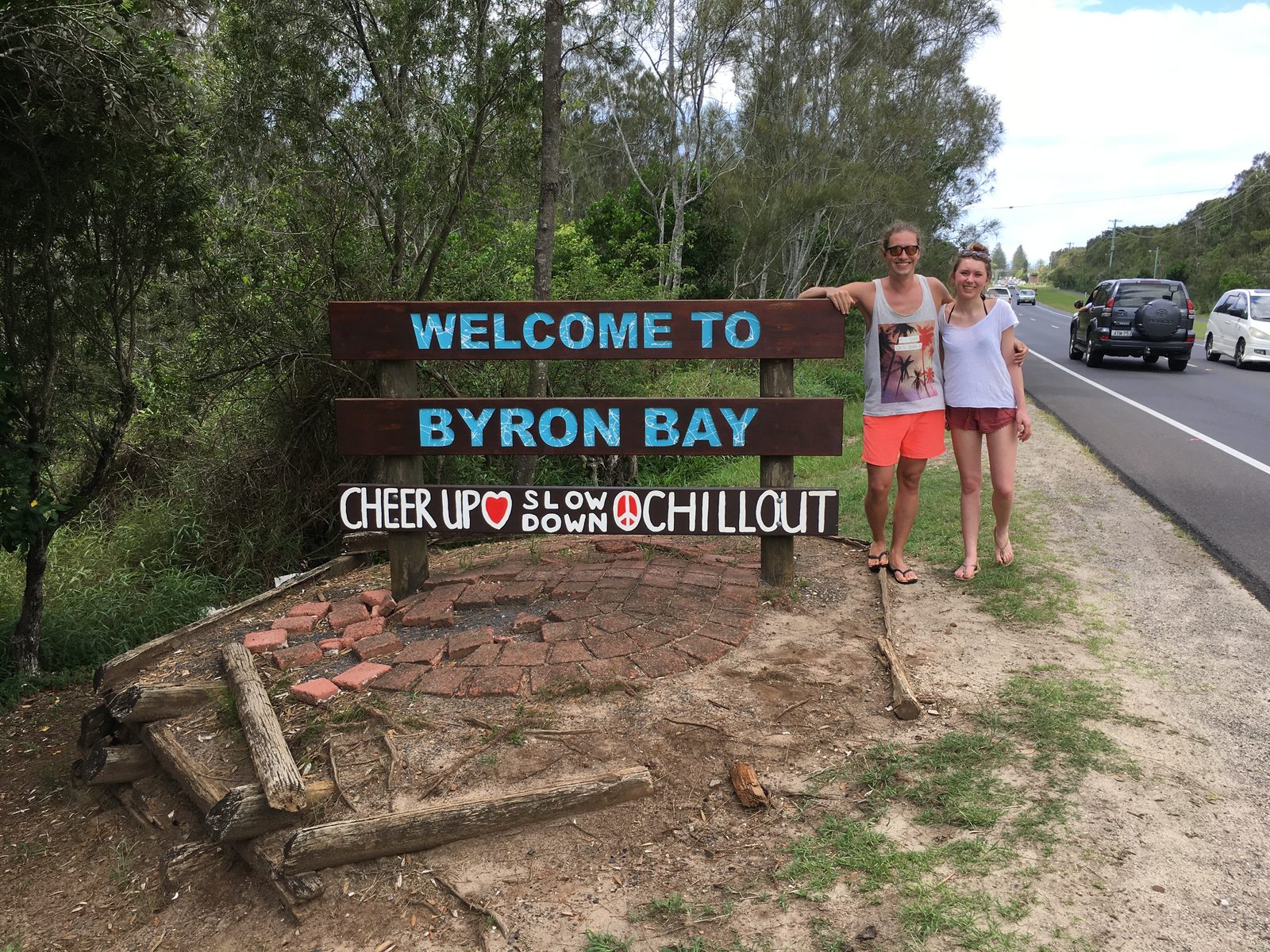 Lucy und Christian stehen an einem Holzschild mit der Aufschrift Welcome to Byron Bay