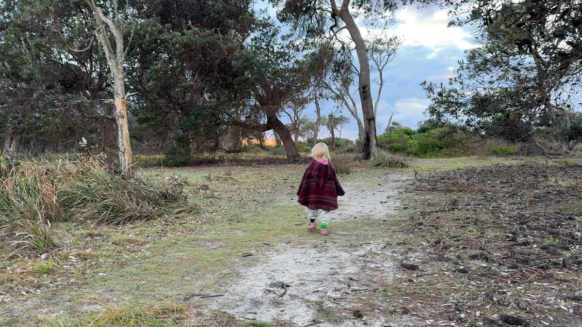 Linnea läuft im roten Poncho allein den sandigen Dünenpfad zwischen Küstenbäumen Richtung Strand, frühes Morgenlicht