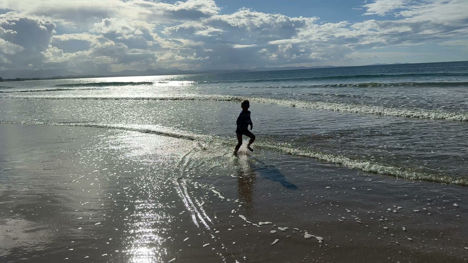 Ein Kind rennt als Silhouette durch die auslaufende Welle am Strand von Byron Bay, das Wasser glitzert im Nachmittagslicht