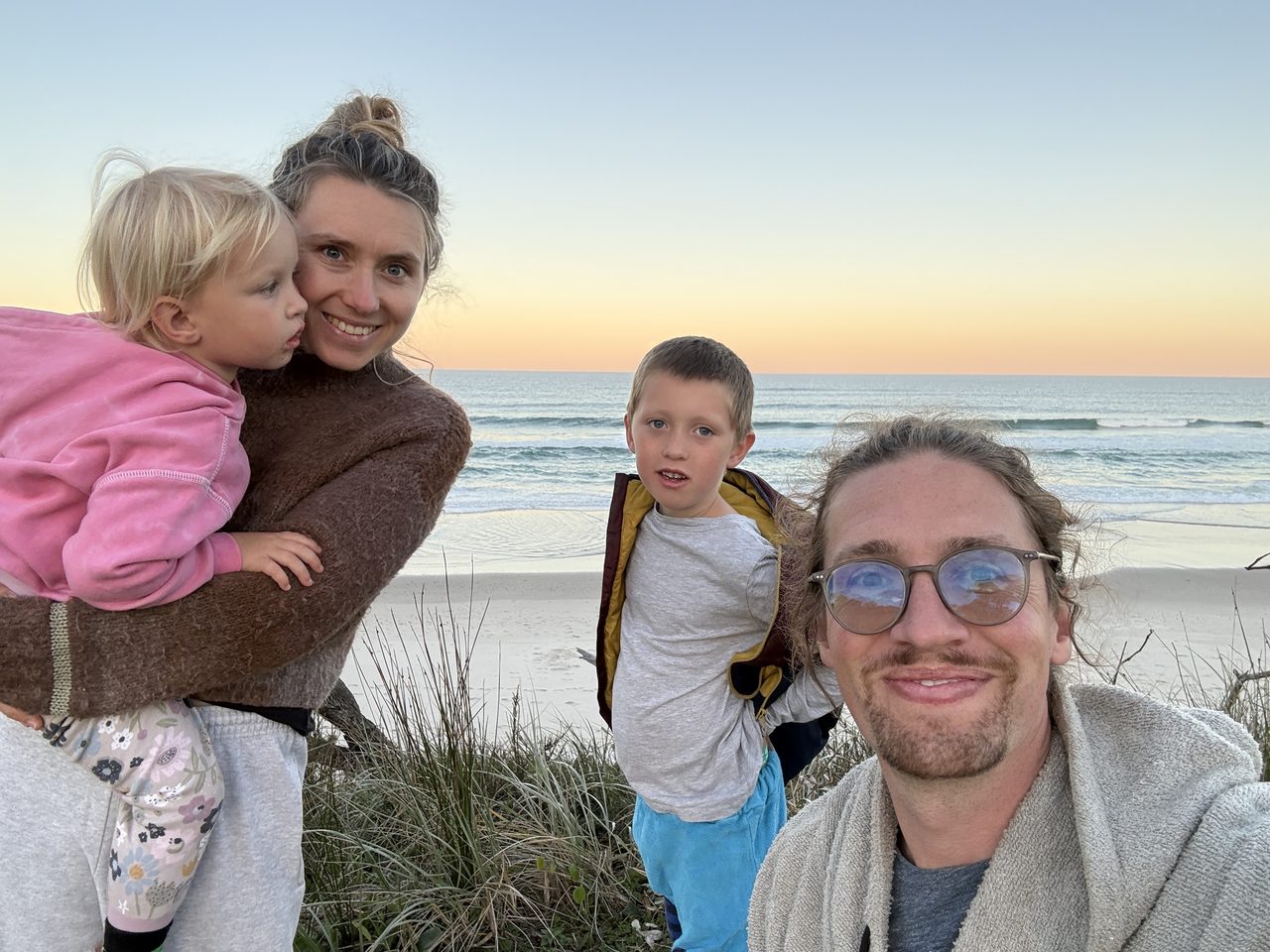 Erstes Familien-Selfie zu viert in Australien: Christian, Lucy mit Linnea auf dem Arm und Joris am Dünenrand von Byron Bay im Abendlicht, dahinter Strand und Brandung