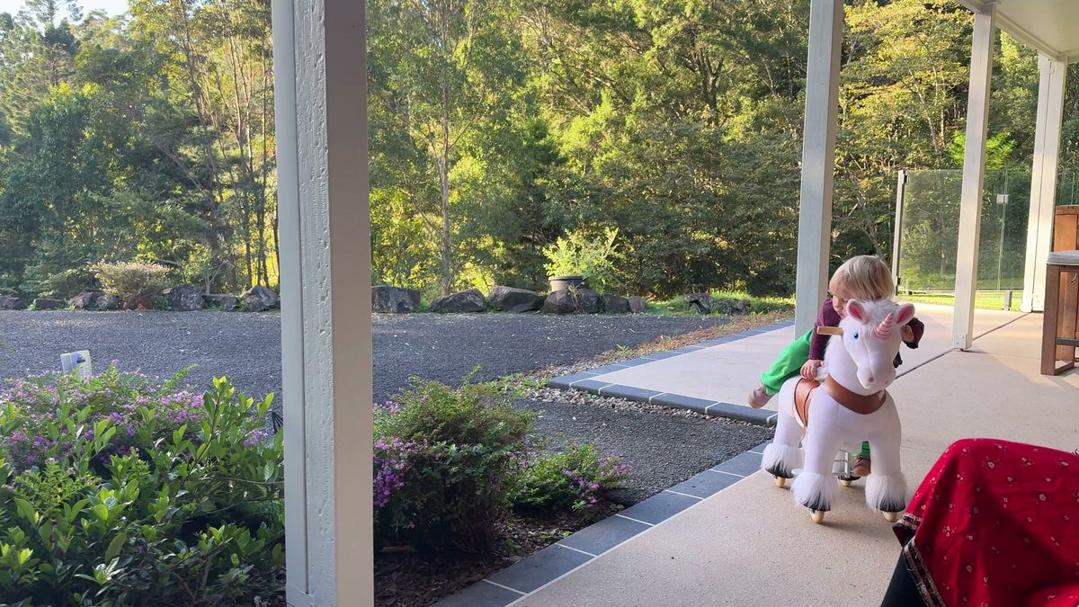 Veranda des Housesits mit weißen Säulen: Linnea sitzt auf ihrem Spielzeug-Einhorn, Joris fährt auf der Kies-Einfahrt Roller, dahinter Regenwald
