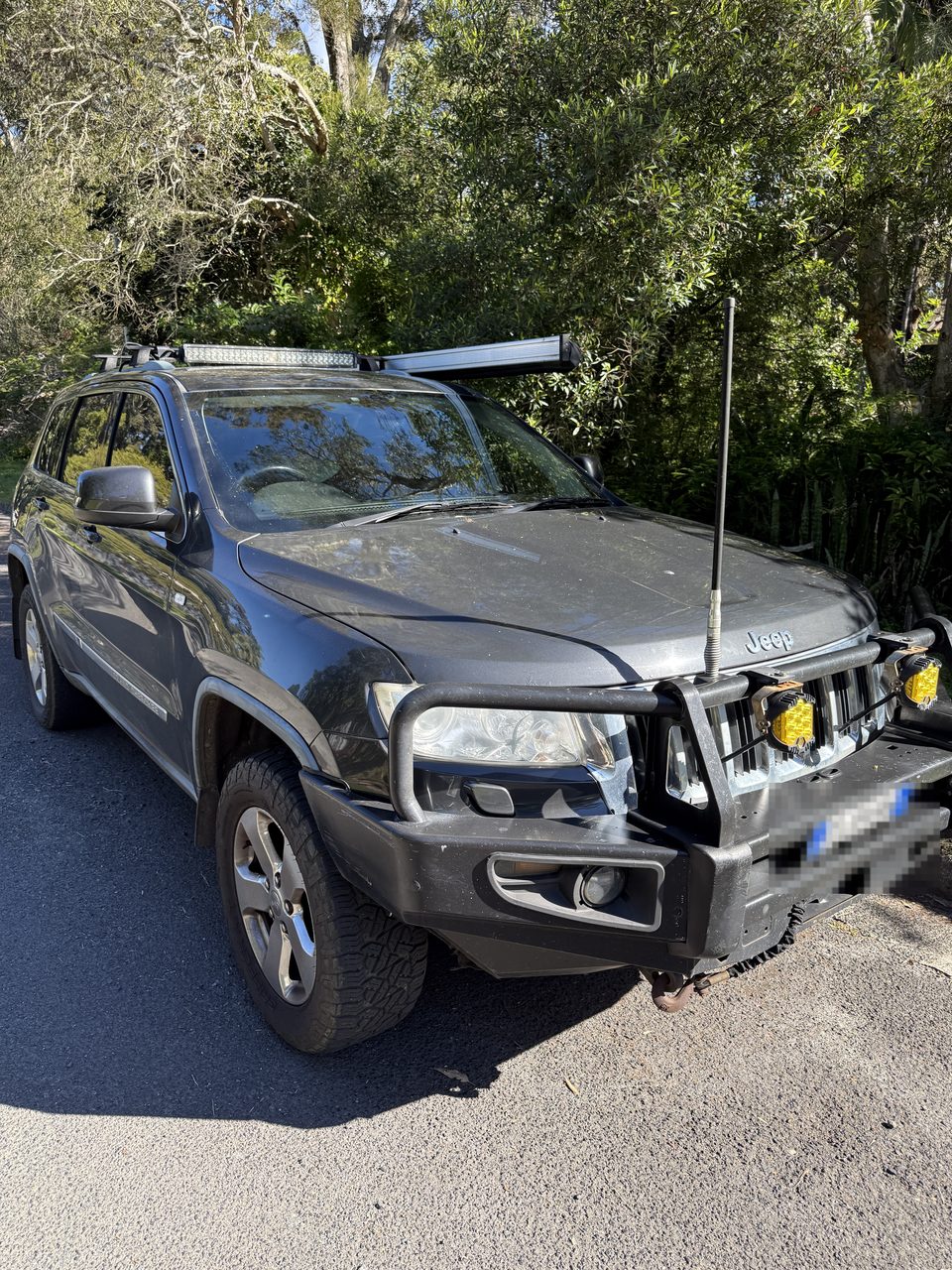 Unser Jeep Grand Cherokee von schräg vorn, mit Dachträger und Lichtbalken, auf einer Straße vor Regenwaldgrün