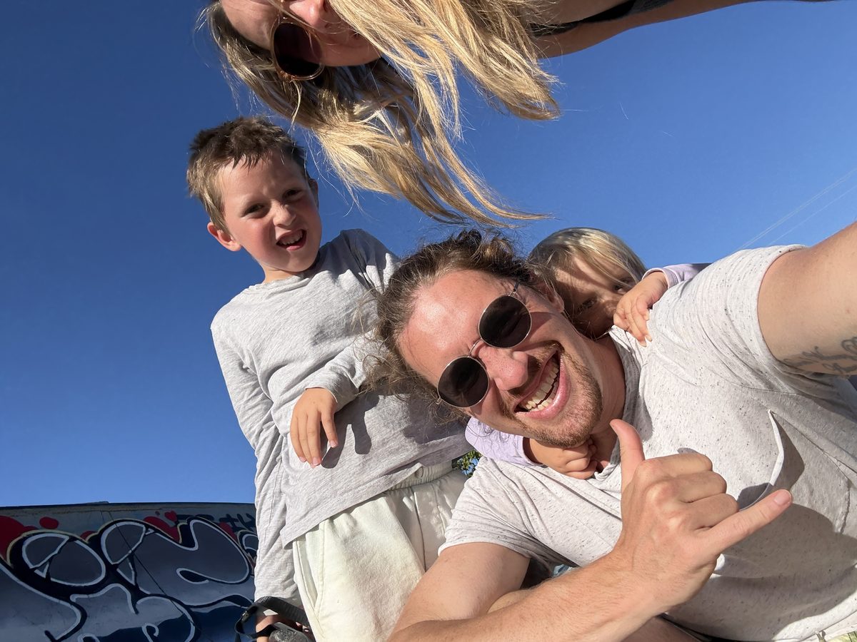 Schiefes Familien-Selfie aus der Froschperspektive im Skatepark: Christian macht die Shaka-Geste, Joris grinst, Lucy und Linnea dahinter, blauer Himmel