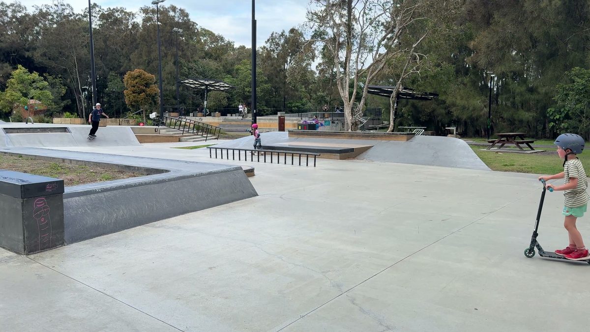 Weitläufiger Betonskatepark zwischen Bäumen, Joris steht mit Helm und Roller am Rand, im Hintergrund weitere Kinder und Jugendliche
