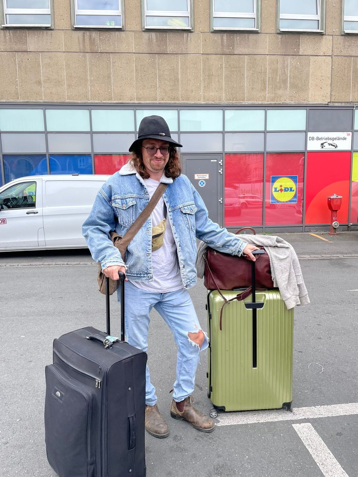 Christian steht mit Koffer, Reisetasche und Trolley allein auf einem Parkplatz vor der Abreise