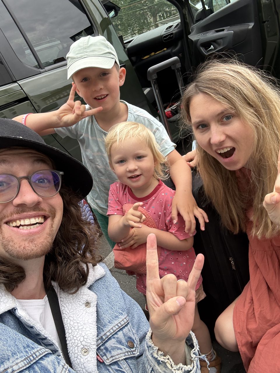 Familien-Selfie am offenen Kofferraum am Abreisetag: Christian mit Hut, Lucy, Joris mit Cap und Linnea, alle lachen in die Kamera